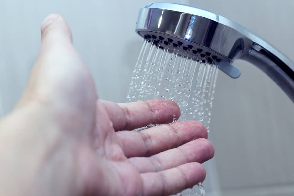 A hand reaching under a running shower to test the water temperature.