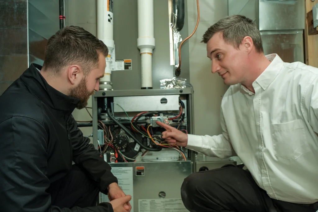 Two technicians are kneeling beside an open gas furnace, with one pointing to the wiring during a diagnostic inspection.
